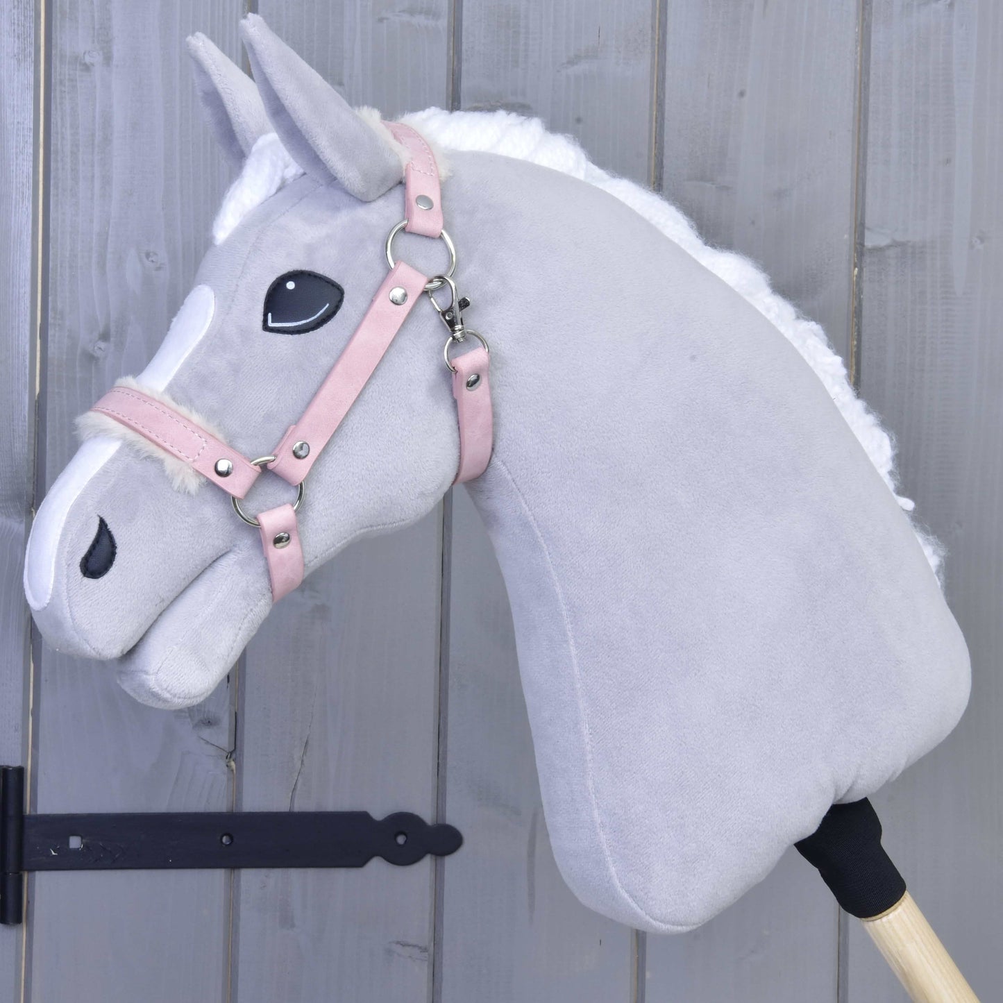 Hobby Horse LarDen Ohlávka s beránkem z koženky Mullbery na světle šedém plyšovém Hobby Horse s bílou hřívou, růžovou uzdečkou z koženky a vyšívanými detaily, nasazeném na dřevěné tyči před šedou dřevěnou stěnou.