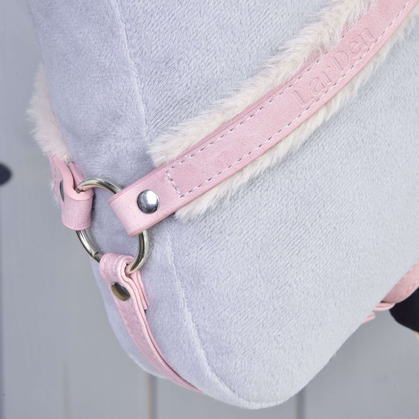 Detail Poprsáku s beránkem Mullbery od Hobby Horse LarDen se světle šedým plyšem, růžovým koženkovým řemínkem s raženým textem, bílým lemem z umělé kožešiny a kovovým kroužkem. Pozadí je rozmazané.