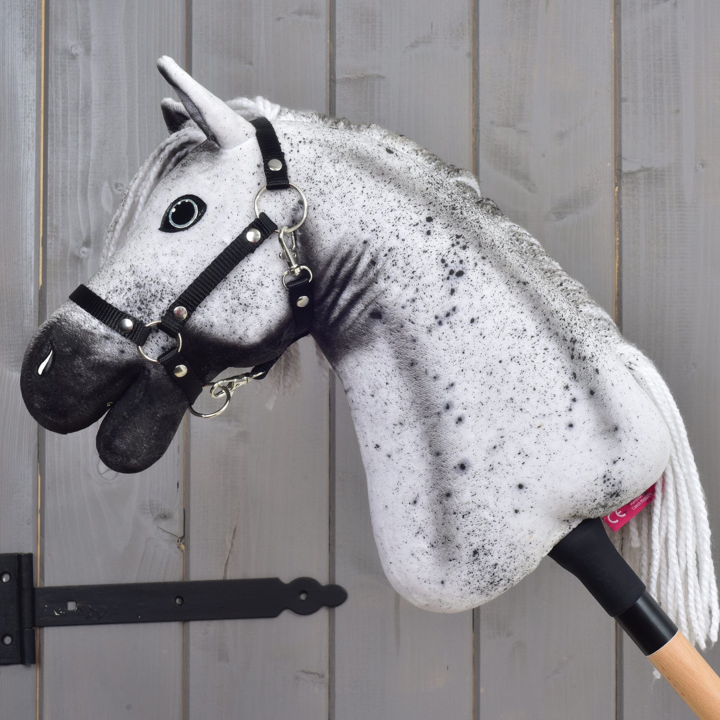 Hobby Horse Naivy s černou ohlávkou - velikost S od Hobby Horse LarDen, s realistickou šedo-černou hlavou hřebce a černou ohlávkou, vystavený před šedou dřevěnou stěnou.