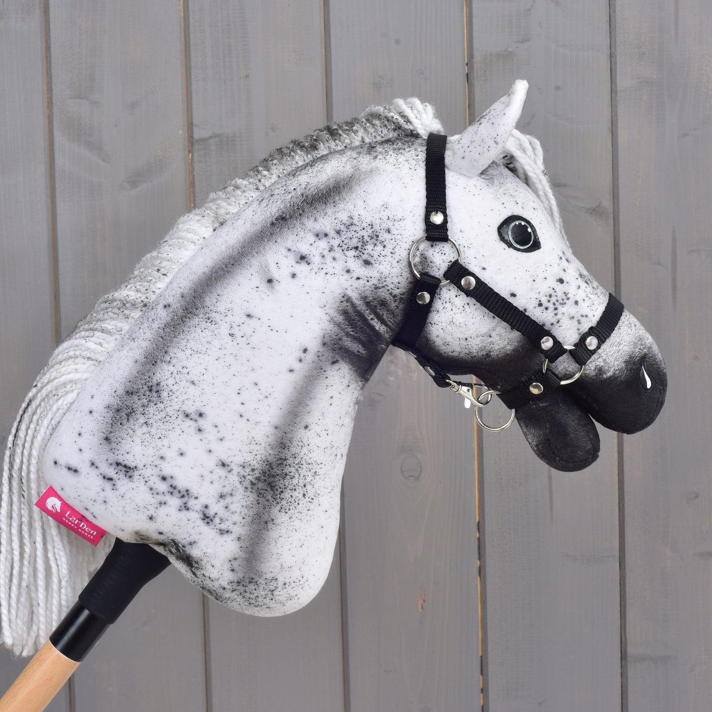 Hobby Horse Naivy s černou ohlávkou - velikost S od Hobby Horse LarDen, s výrazným bílo-šedým skvrnitým vzhledem, hrdě stojí nasazený na dřevěné tyči před šedou dřevěnou stěnou.
