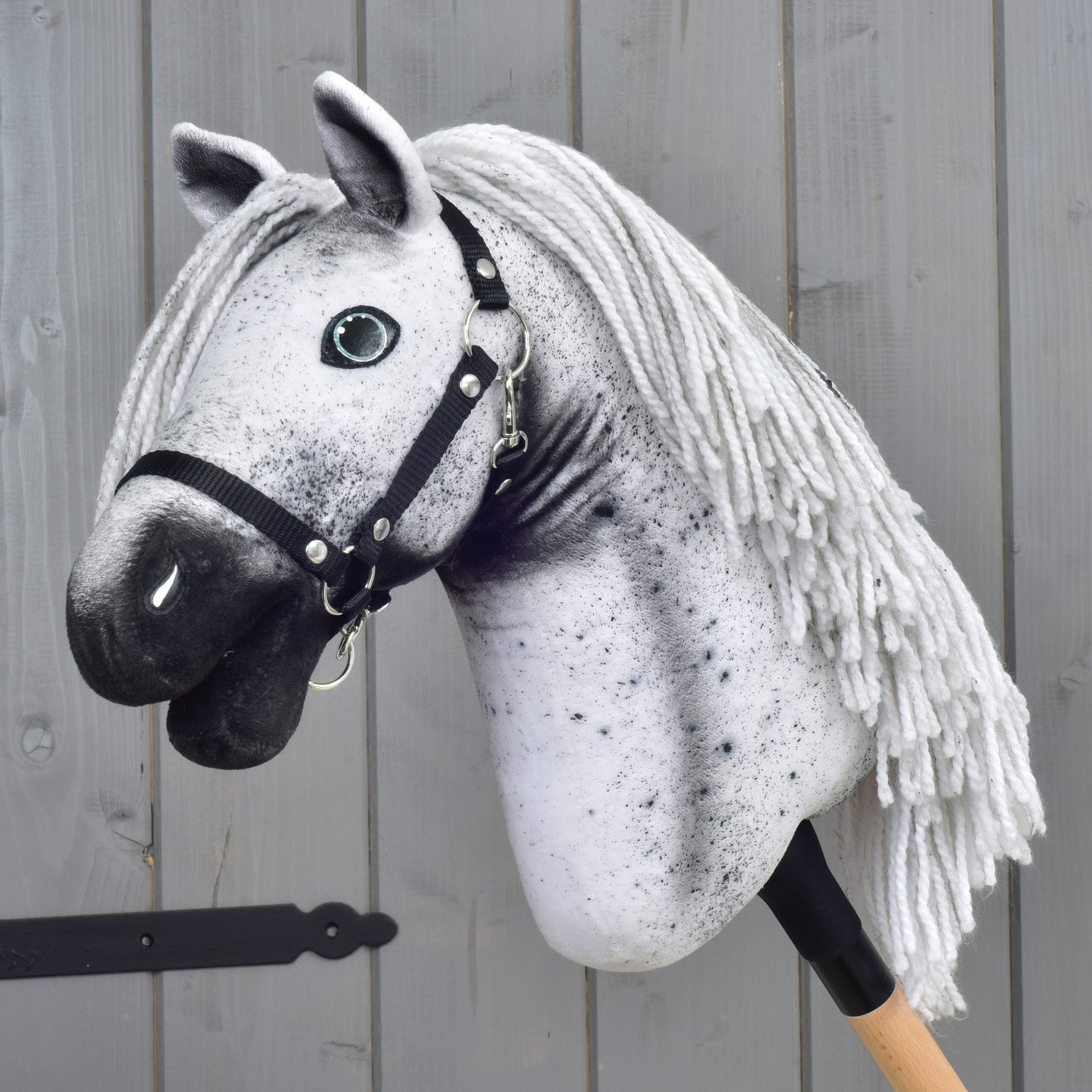 Hobby Horse LarDen "Hobby Horse Naivy with Black Halter" (velikost S), se šedo-bílým appaloosa vzhledem, černou ohlávkou a přízovou hřívou, nasazený na dřevěné tyči a umístěný před šedou dřevěnou stěnou.