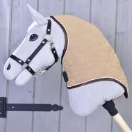 Nákrční díl Bonanza od Hobby Horse LarDen, detail šedého Hobby Horse s černou uzdečkou a béžovou prošívanou dekou s bezpečným zapínáním, nasazeného na dřevěné tyči před šedou dřevěnou stěnou.