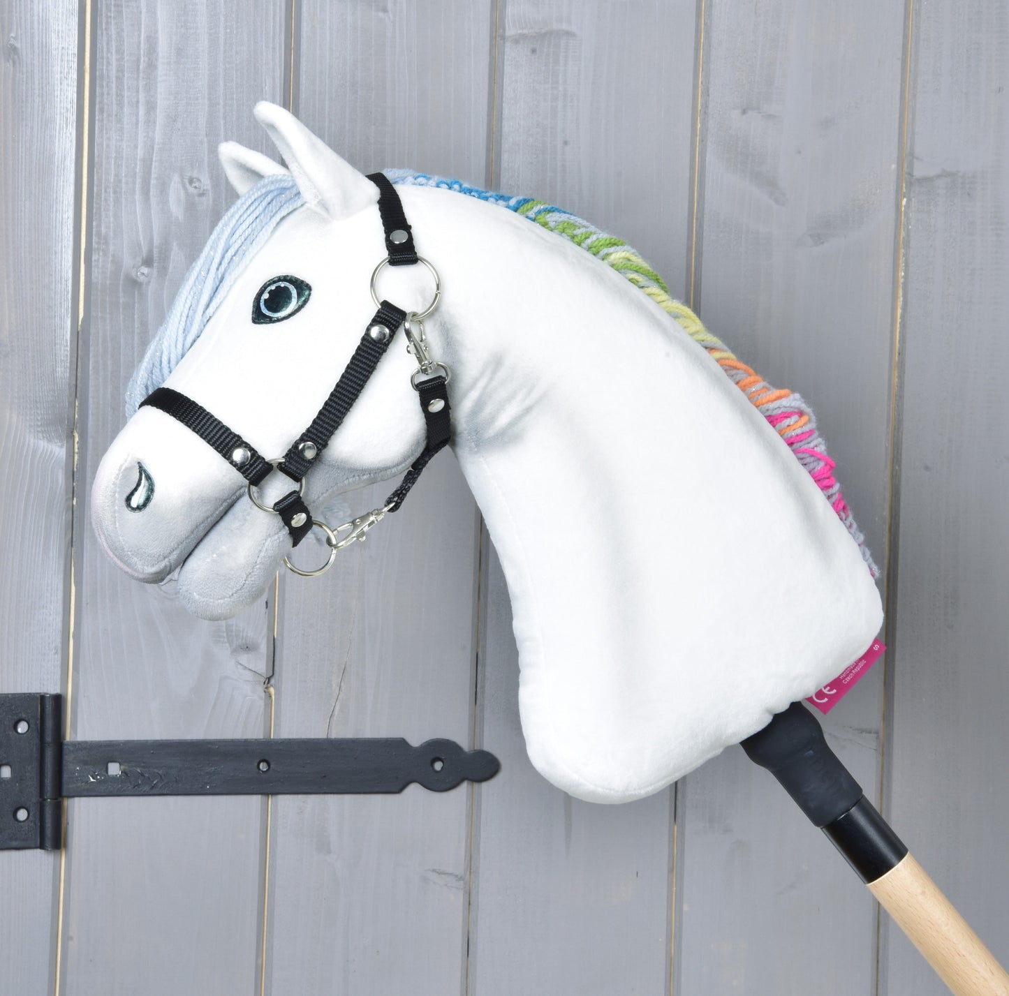 Hobby Horse Rainbow New od Hobby Horse LarDen, bílý hračkový kůň s černou uzdečkou a duhovou hřívou na dřevěné tyči, opřený o šedou dřevěnou stěnu.