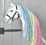 Hobby Horse LarDen „Hobby Horse Rainbow New“ má bílý obličej, černou uzdečku a dlouhou duhově zbarvenou hřívu z příze, stojí před šedými dřevěnými dveřmi.