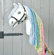 Hobby Horse Rainbow New od Hobby Horse LarDen má ručně vyrobený bílý obličej, modré oči, černou uzdečku a duhovou hřívu z modré, zelené, žluté a růžové příze na světle šedém dřevěném pozadí pro pohádkový dojem.