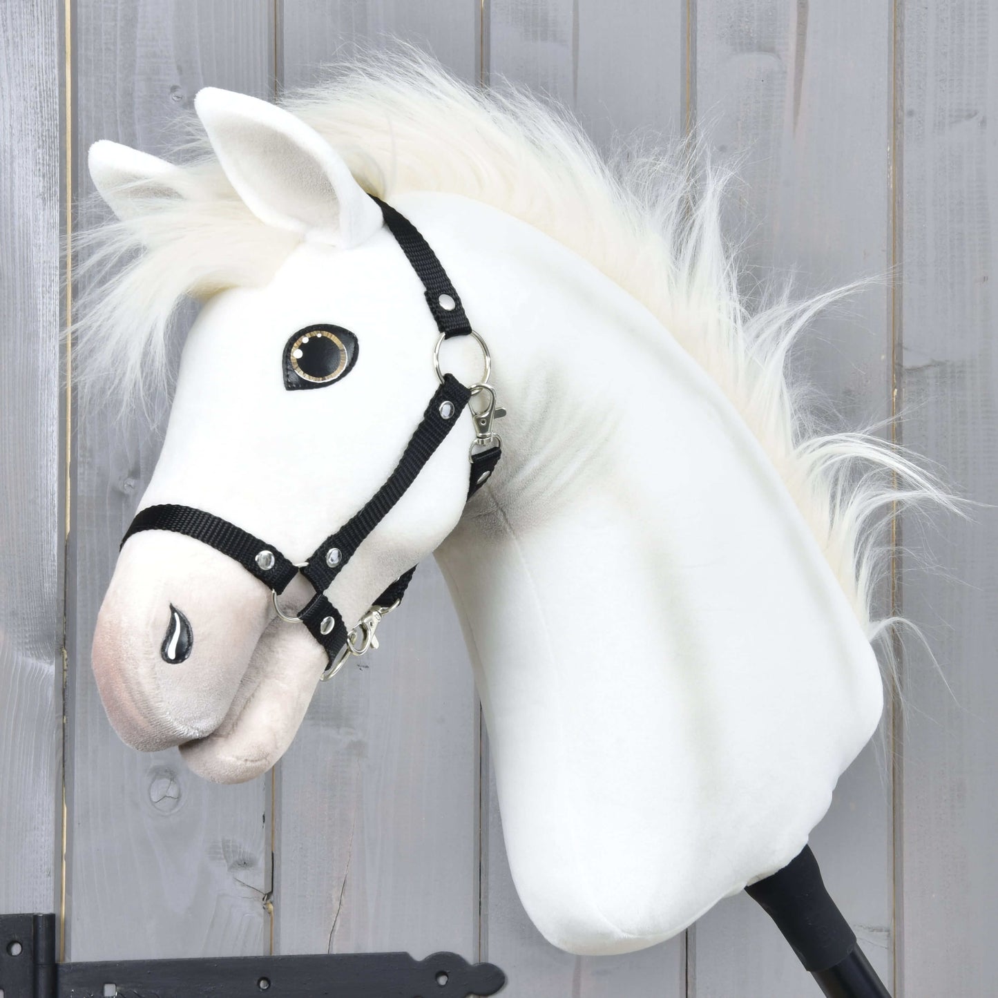 Hobby Horse Nexus s černou ohlávkou od Hobby Horse LarDen, s bílou plyšovou hlavou inspirovanou plemenem Akhal-Teke, knoflíkovým okem a huňatou hřívou, zobrazený na pozadí dřevěné stěny – ideální pro každého nadšence do Hobby Horse Nexus.