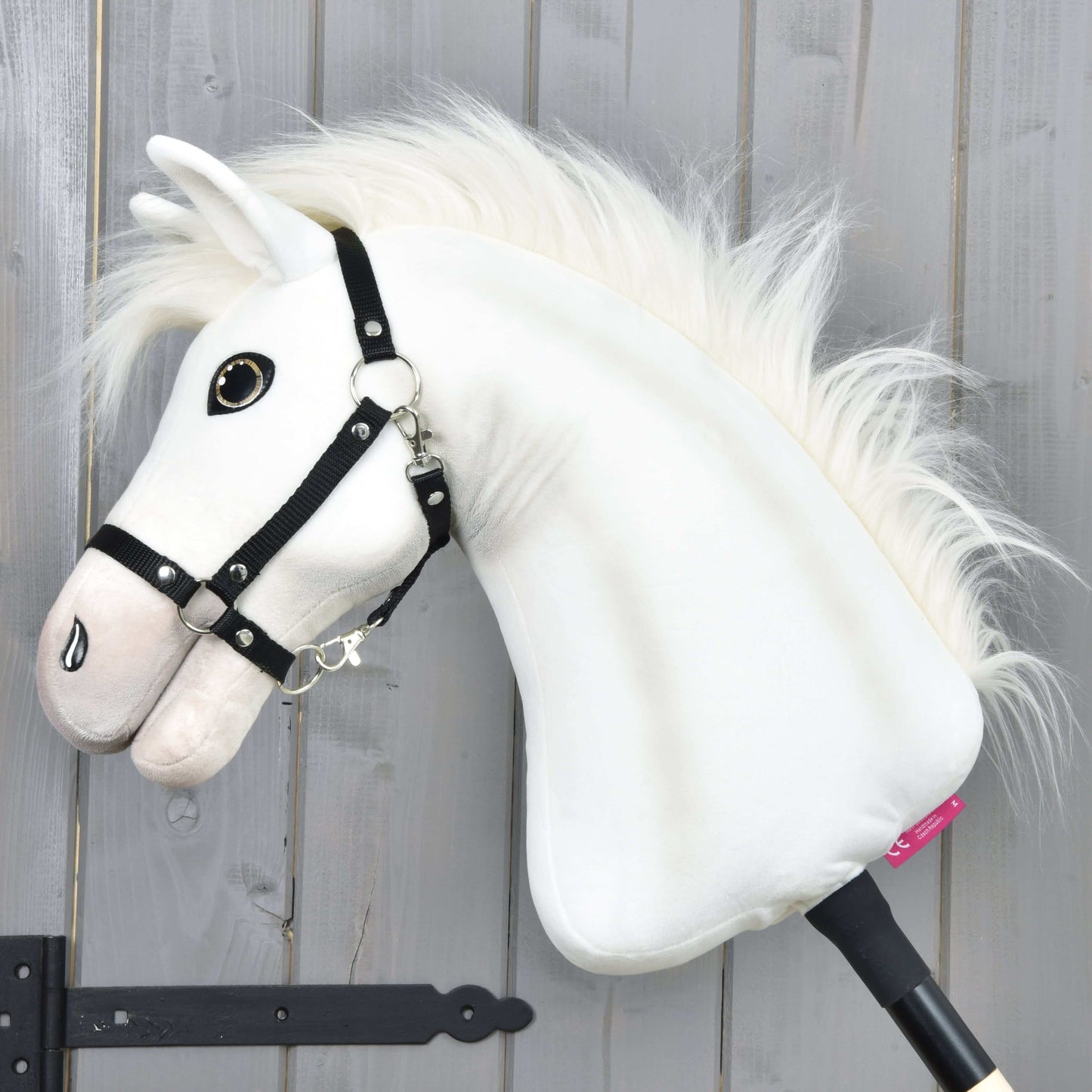 Hobby Horse Nexus s černou ohlávkou od Hobby Horse LarDen, bílý plyšový sportovní kůň s huňatou hřívou, velkým knoflíkovým okem a černou ohlávkou, vyfotografovaný před šedou dřevěnou stěnou – ideální pro každého nadšence.
