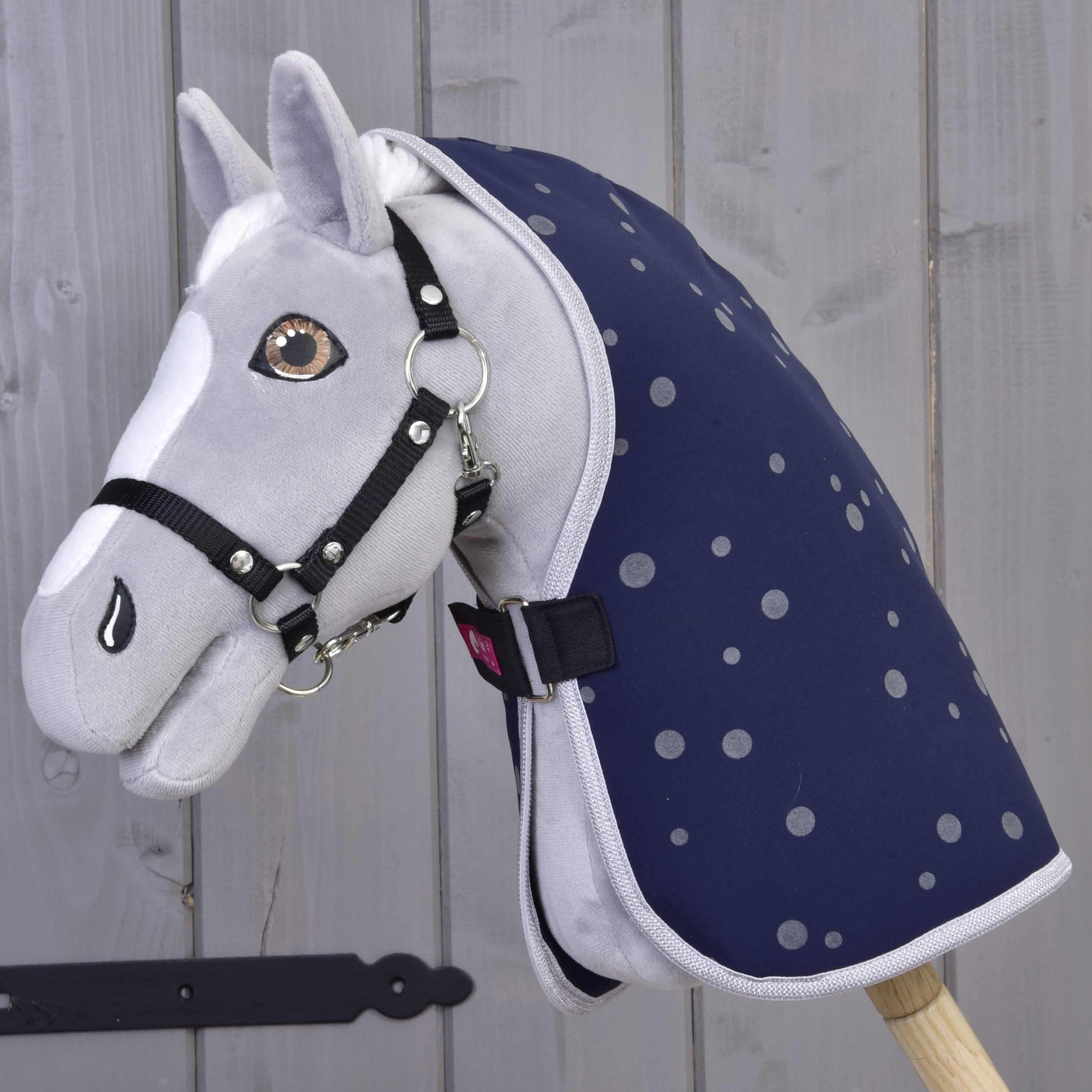 Šedý plyšový Hobby Horse od Hobby Horse LarDen s černou uzdečkou a Dekou stájovou Night Sky, nasazený na dřevěné tyči na pozadí světlé dřevěné stěny.