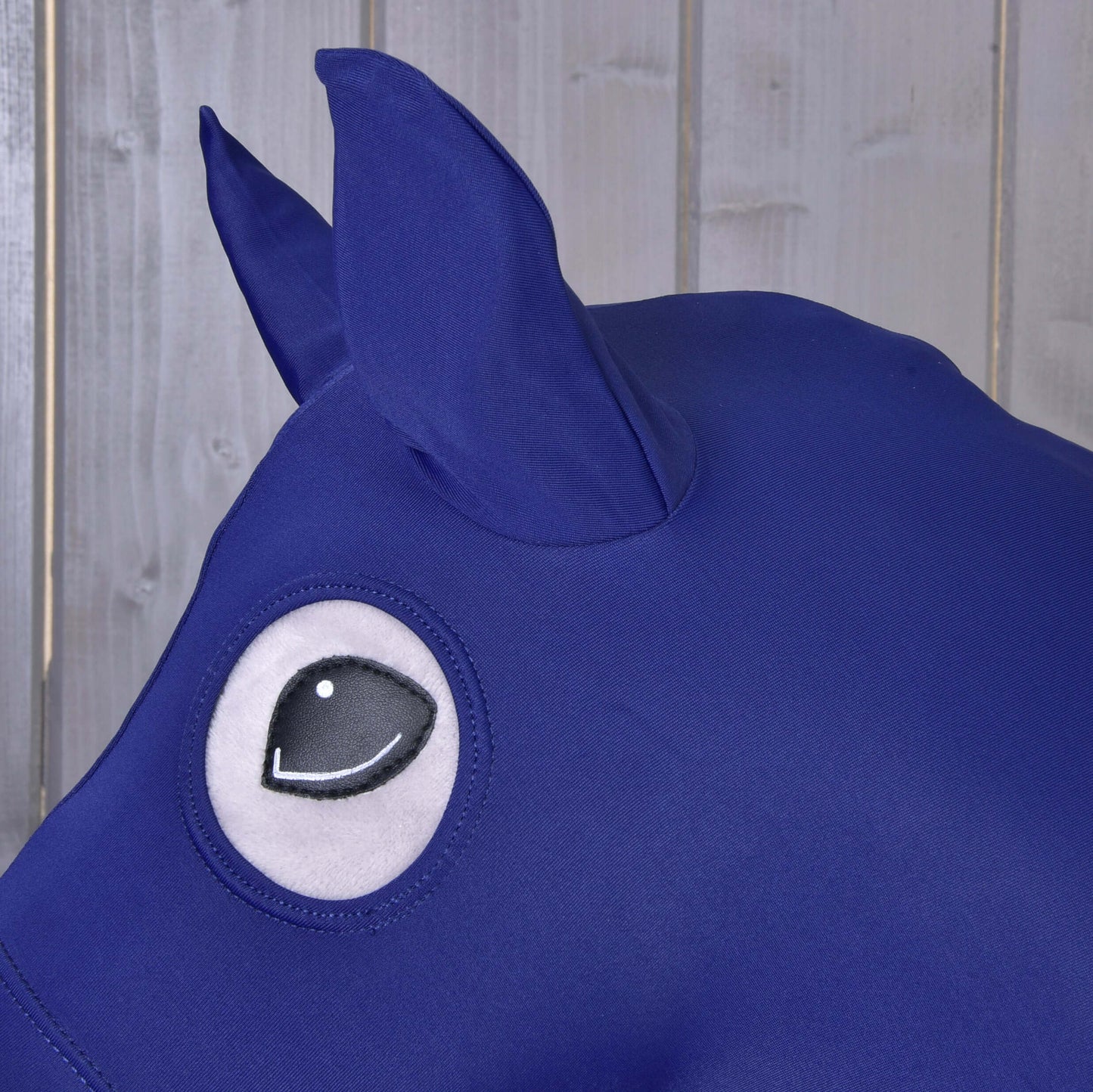 Detail modrého ochranného obleku Hobby Horse LarDen z Lycry, navrženého pro Hobby Horse, s velkým okem v kresleném stylu a špičatým uchem, zobrazeného na pozadí světlých dřevěných panelů.