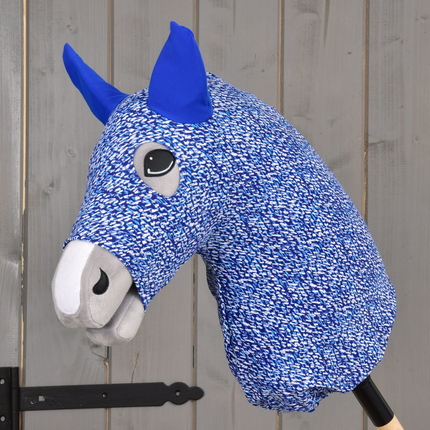 Detail Hobby Horse ochranného obleku See od Hobby Horse LarDen s modro-bílou PES látkou, modrýma ušima a šedým čenichem, nasazeného na dřevěné tyči před šedou dřevěnou stěnou.