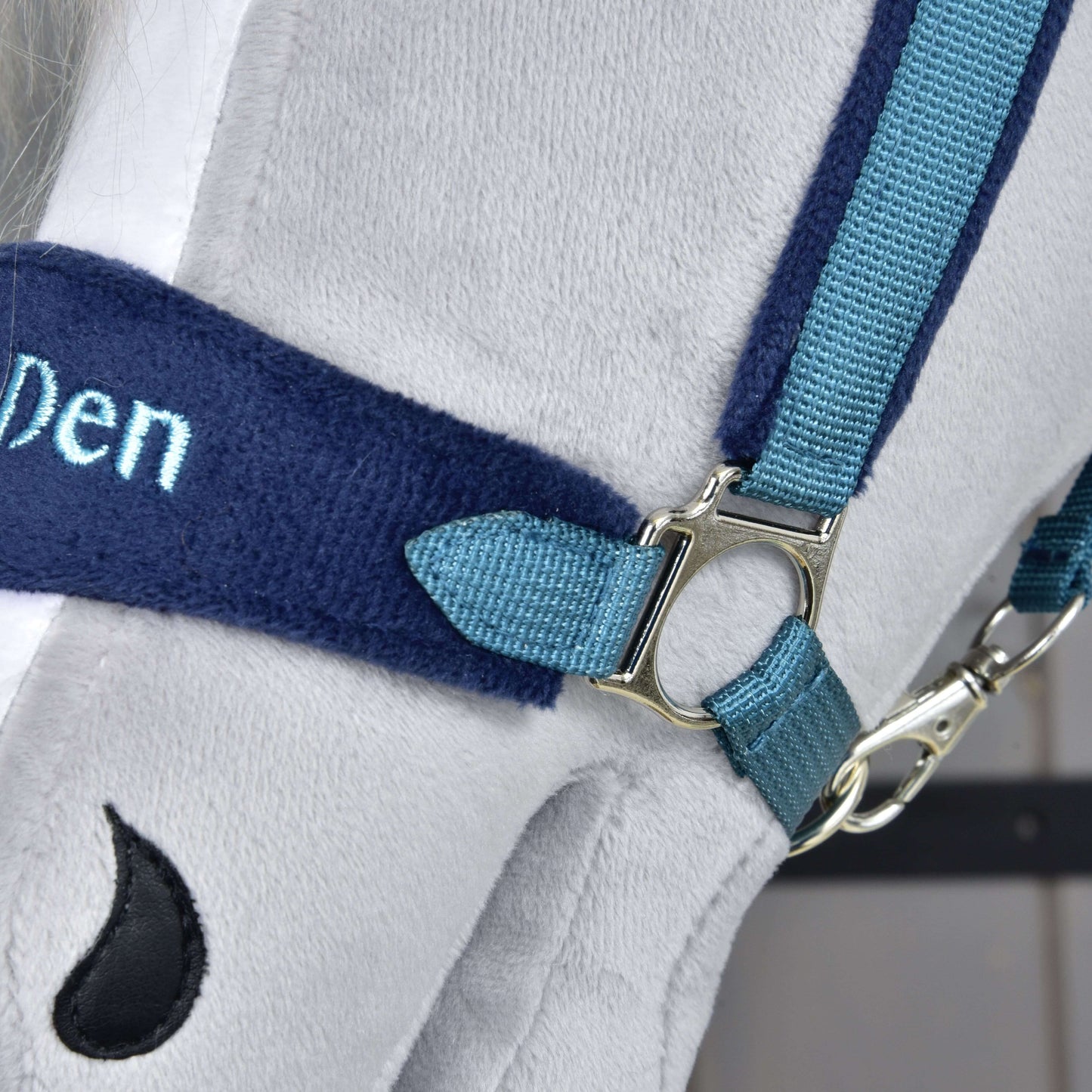 Ohlávka s beránkem LarDen Blue od Hobby Horse LarDen, detail měkké šedé látky, modrých popruhů, stříbrné přezky, částečně viditelné výšivky „Den“ a malého černého detailu ve tvaru kapky. Perfektní jako stylový doplněk ohlávky pro Hobby Horse.