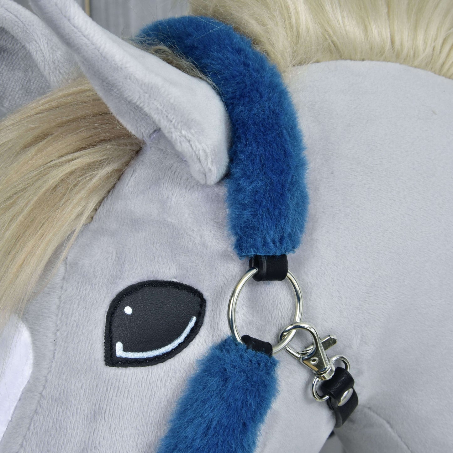 Detail hlavy šedého plyšového koně s krémovou hřívou, černobíle vyšitým okem a ohlávkou Hobby Horse LarDen Ohlávka s beránkem Teddy Petrol v modré barvě s kovovými kroužky a zapínáním.