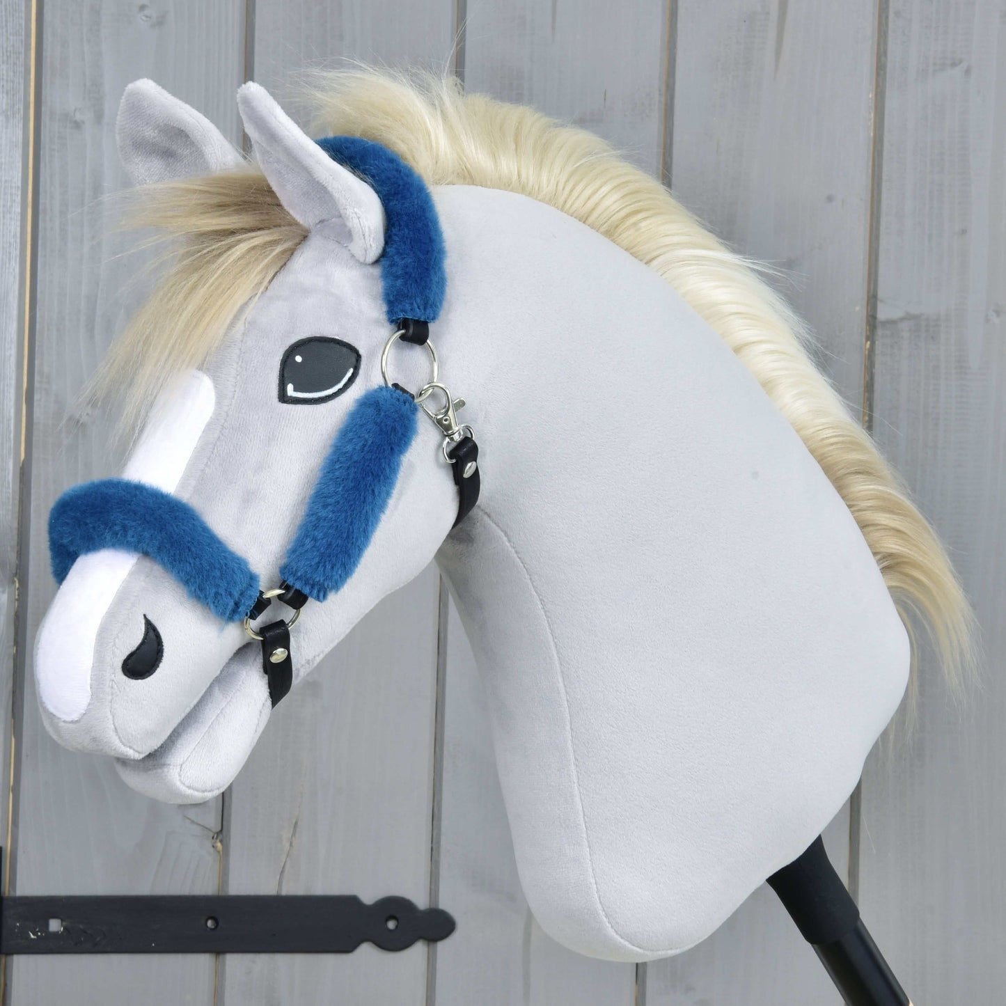 Hobby Horse LarDen Ohlávka s beránkem Teddy Petrol na plyšovém Hobby Horse se světle šedou látkou, bílým mulem, blond hřívou a modrou koženkovou ohlávkou na černé tyči, před dřevěným pozadím.