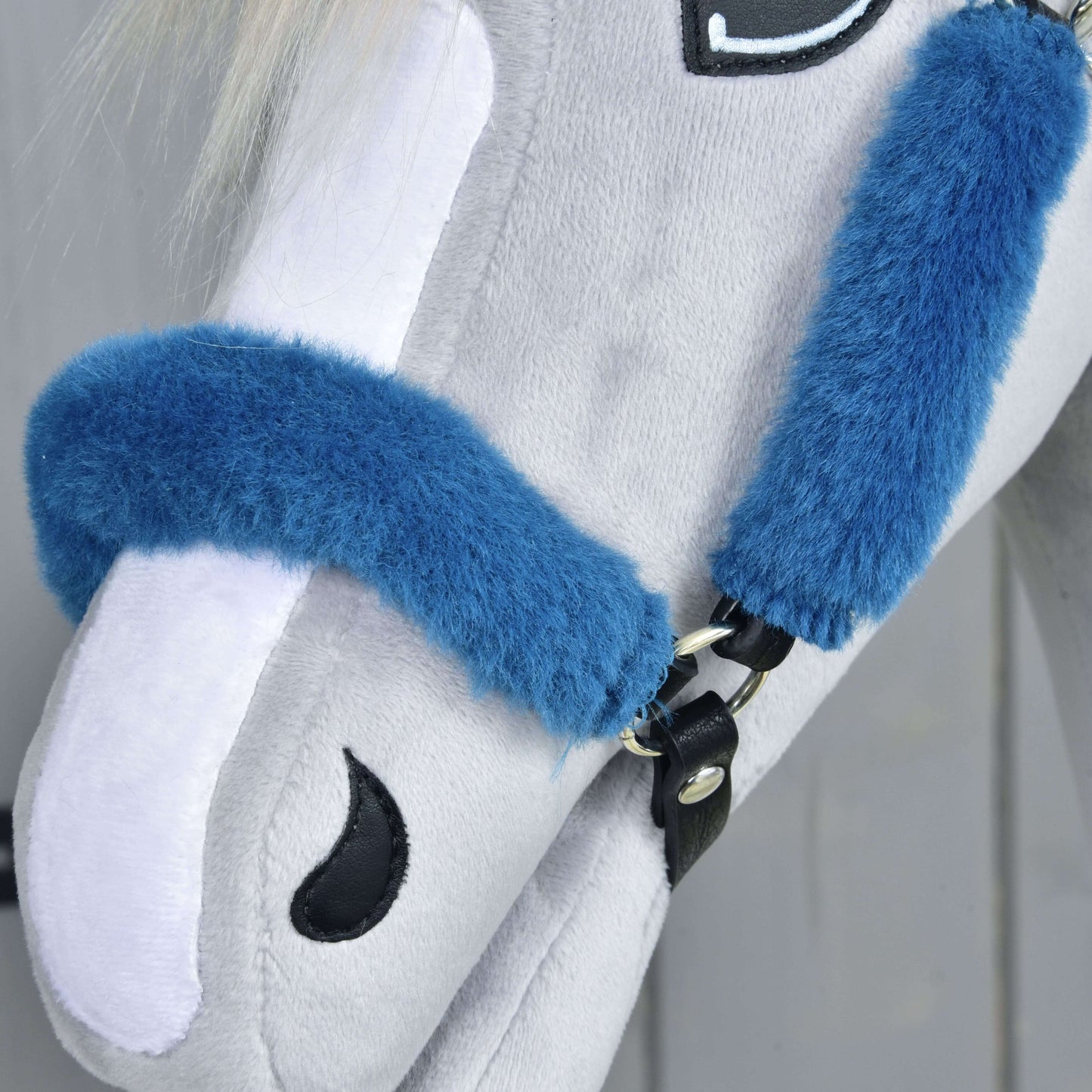 Detail hlavy světle šedého plyšového koně s ohlávkou s beránkem Teddy Petrol od Hobby Horse LarDen, s modrou ohlávkou, bílou hřívou a vyšívanými černými detaily oka a nozder na jemně rozostřeném neutrálním pozadí.