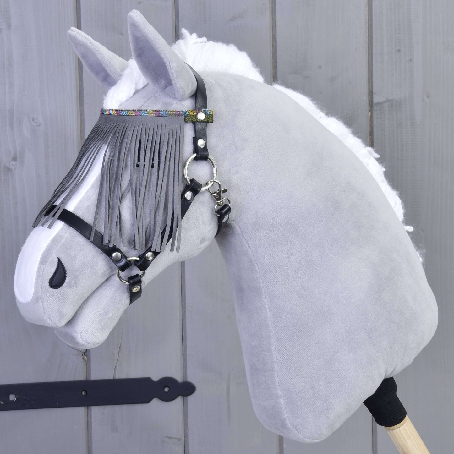 Plyšová šedá hlava Hobby Horse s bílou hřívou, s ohlávkou od Hobby Horse LarDen v černé barvě se šedými třásněmi a šedým nánosníkem, na dřevěné tyči, před dřevěnou stěnou.