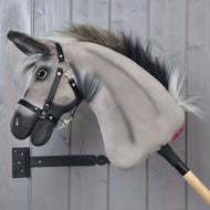 Hobby Horse osel Olin s černou ohlávkou od Hobby Horse LarDen se šedou plyšovou oslí hlavou, tmavou hřívou, černou ohlávkou, otěžemi a stříbrným kroužkem u tlamy na dřevěné tyči, před šedou zdí.