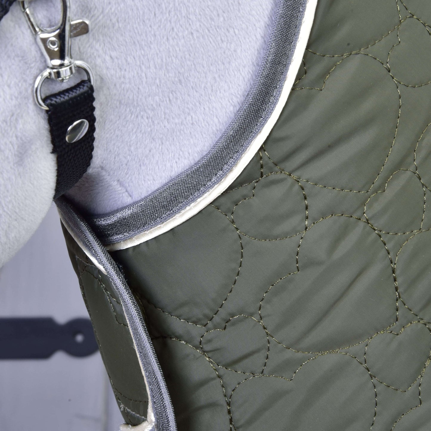 Detail zelené prošívané podsedlové dečky Set Olivetti s bílým lemováním od Hobby Horse LarDen, s kovovou sponou na černém popruhu, pevně nasazené na světle šedém koni.