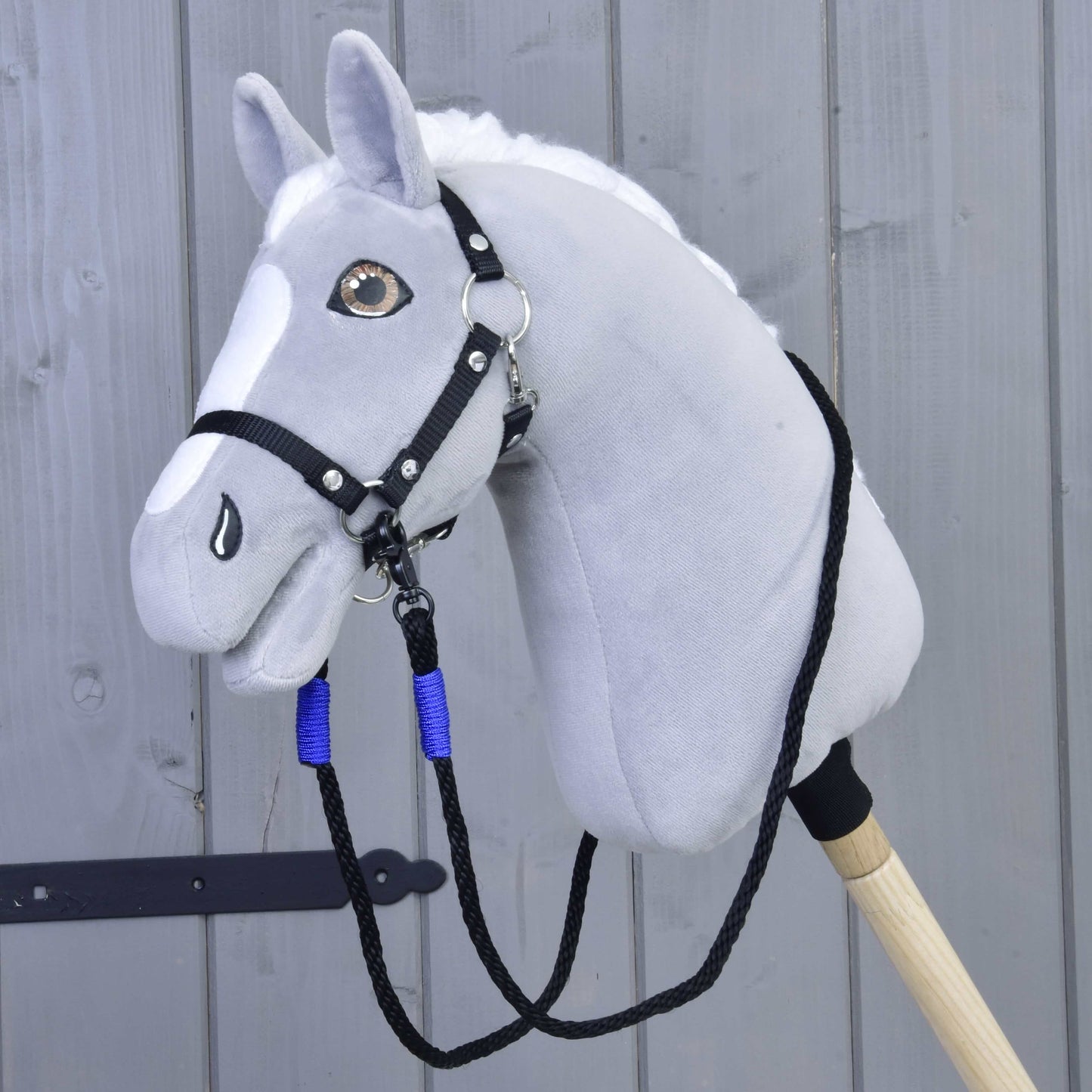 Otěže Basic Blue od Hobby Horse LarDen zobrazené na šedé plyšové hlavě Hobby Horse s černou uzdečkou a modrými polypropylenovými provazovými otěžemi na dřevěné tyči, na pozadí světle šedé dřevěné stěny.
