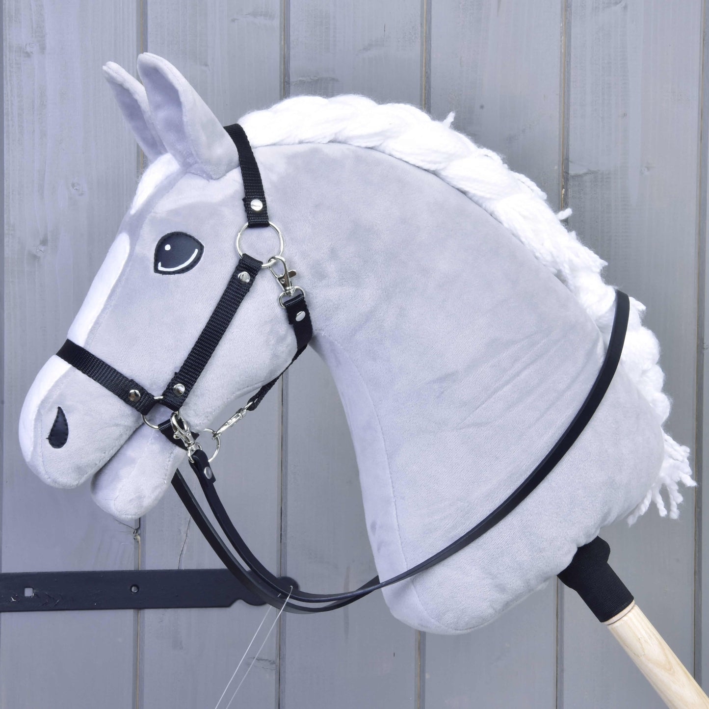 Černé otěže z koženky od Hobby Horse LarDen na šedém plyšovém Hobby Horse s bílou hřívou, bezudidlovou uzdečkou a kvalitními černými otěžemi z koženky, na dřevěné tyči před šedým dřevěným panelovým pozadím.