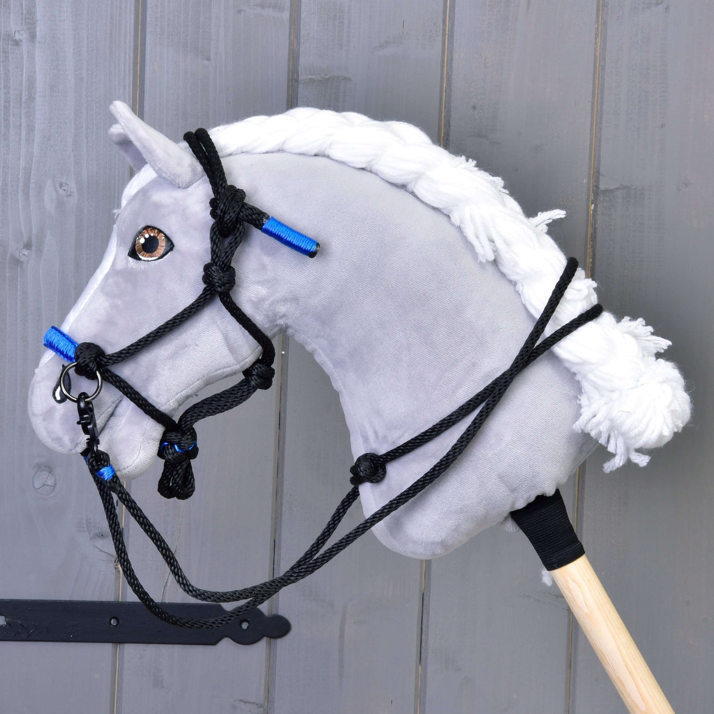 Parelka Azure od Hobby Horse LarDen na plyšovém šedém Hobby Horse s bílou přízovou hřívou, černou uzdečkou a výraznými azurovými akcenty, nasazeném na dřevěné tyči před šedou dřevěnou stěnou.