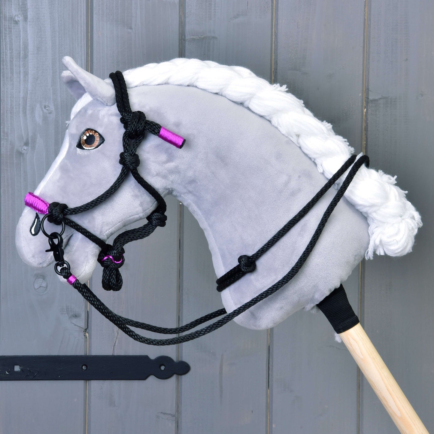 Parelka Violette od Hobby Horse LarDen na šedém Hobby Horse s bílou hřívou, černou uzdečkou a detaily otěží, fialovými akcenty a stylovou parelkou, nasazeném na dřevěné tyči před šedou dřevěnou stěnou.