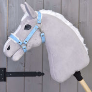 Šedý plyšový koník na tyči s bílou hřívou má na sobě Hobby Horse LarDen Ohlávka s beránkem z koženky Pastell Blue a je opřený o dřevěnou panelovou stěnu s černými kovovými panty.