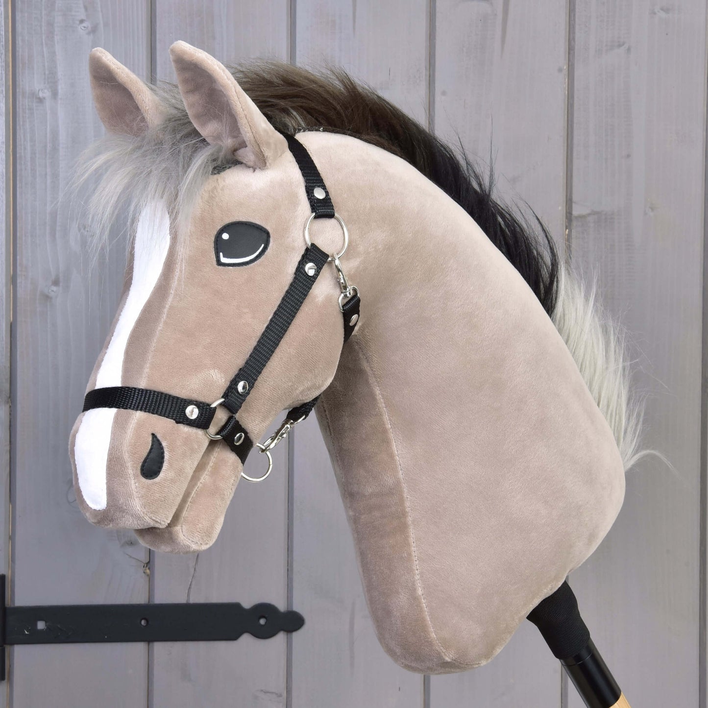 Hobby Horse Piccolino s černou ohlávkou od Hobby Horse LarDen, světle hnědý plyšový sportovní kůň plemene holštýn s bílým pruhem na hlavě, černou ohlávkou, huňatou hřívou a na dřevěné tyči, před šedou dřevěnou stěnou.