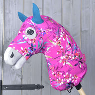 Hobby Horse ochranný oblek Pink Beauty (Lycra) od Hobby Horse LarDen s výraznou růžovou květovanou látkou, modrýma ušima a bílým čenichem, nasazený na dřevěné tyči před šedou dřevěnou stěnou.
