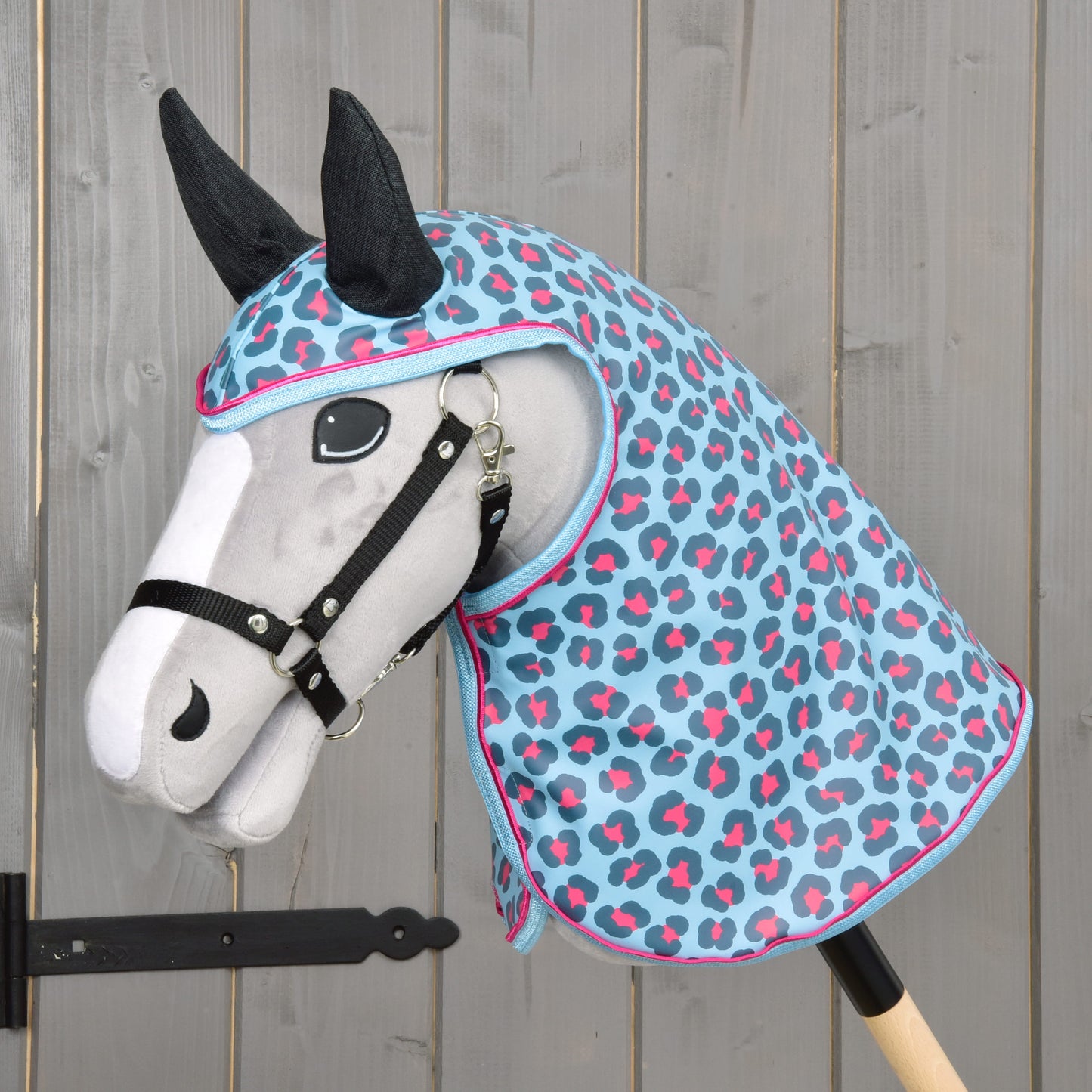 Hobby Horse LarDen Pláštěnka Blue Leopard s plyšovým Hobby Horse s bílým obličejem, černou uzdečkou a ušima a modrou, růžovou a šedou kapucí pláštěnky se skvrnitým vzorem – vše na pozadí šedého dřeva.