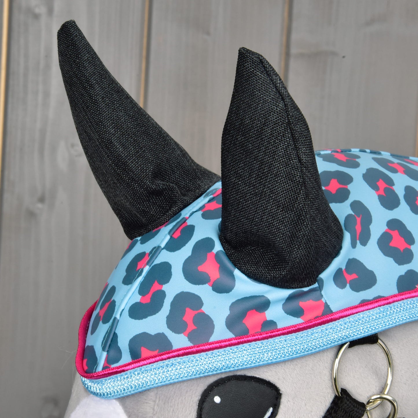 Detail plyšové hračky Hobby Horse LarDen „Raincoat Blue Leopard“ v modro-růžové čepici s leopardím vzorem a černými rohy, připravené na dobrodružství v jakémkoli počasí, na dřevěném pozadí.