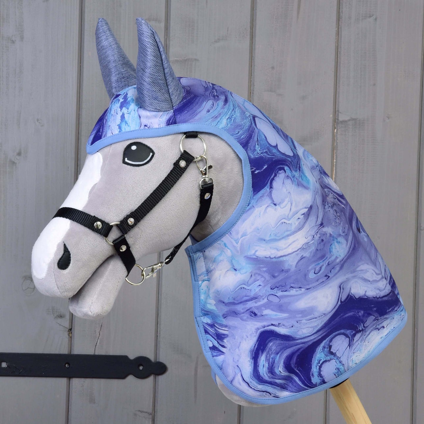 Hobby Horse LarDen „Pláštěnka Galaxy Blue“ s plyšovým koněm, černou uzdečkou a kapucí s modrým, fialovým a bílým mramorovaným vzorem, zobrazený před šedou dřevěnou stěnou.