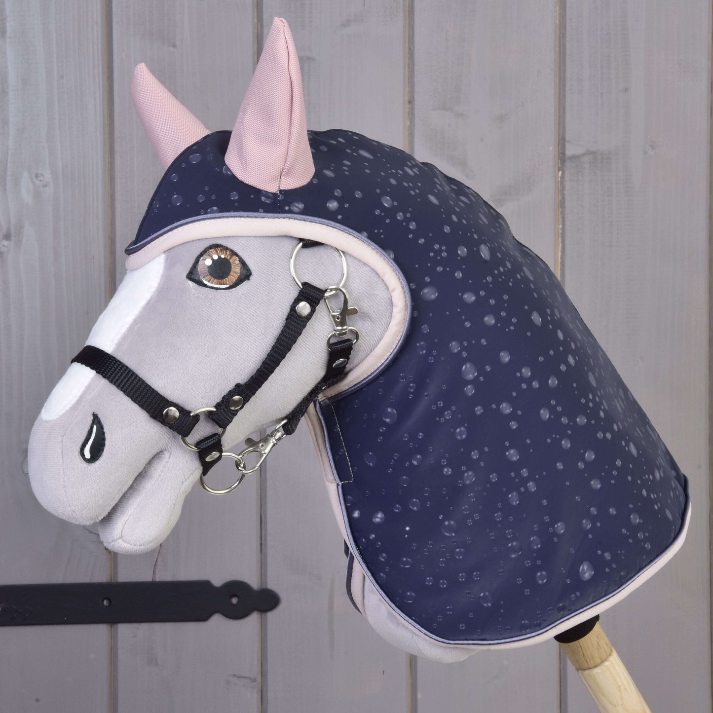 Hobby Horse LarDen Pláštěnka Pinkie Blue (softshell) s šedo-bílým Hobby Horse s růžovými ušima a tmavě modrou pláštěnkou se vzorem deště, černou uzdečkou, perfektní pro hru za každého počasí, zobrazeno na pozadí světlého dřevěného obložení.