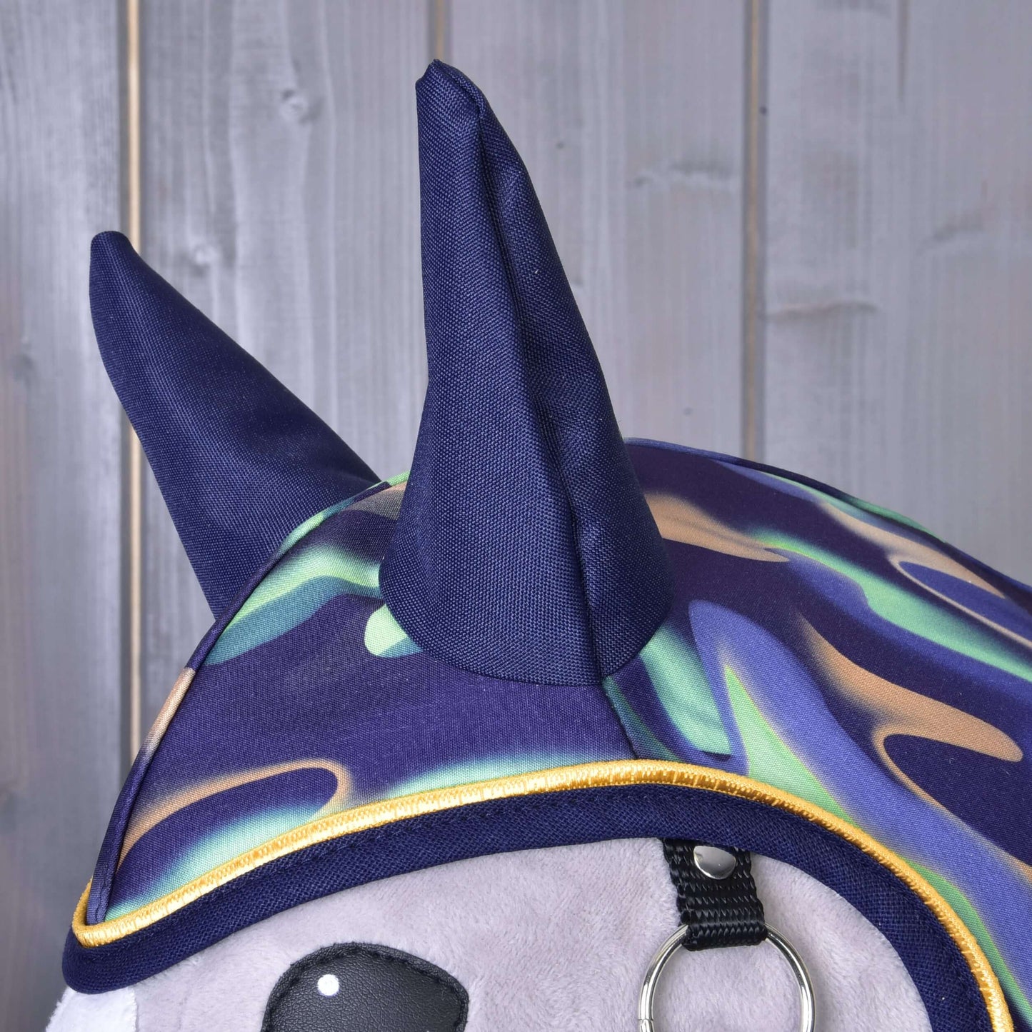 Detail Pláštěnky Rainbow od Hobby Horse LarDen: plyšová hračka v barevné batikované kapuci se dvěma tmavě modrými rohy, stylizované jako hravá nepromokavá pláštěnka, na pozadí ze světle šedého dřevěného obložení.