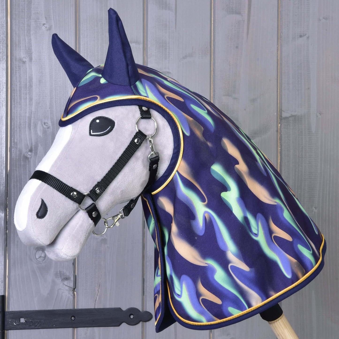 Plyšový Hobby Horse s černou uzdečkou má na sobě Pláštěnku Rainbow od Hobby Horse LarDen s lesklým vícebarevným designem přes hlavu a krk, na dřevěném pozadí.