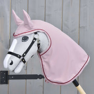 Šedý Hobby Horse LarDen na dřevěné tyči má černou uzdečku a přes hlavu a krk růžovou Pláštěnku Veloria, na pozadí světlého dřevěného plotu.