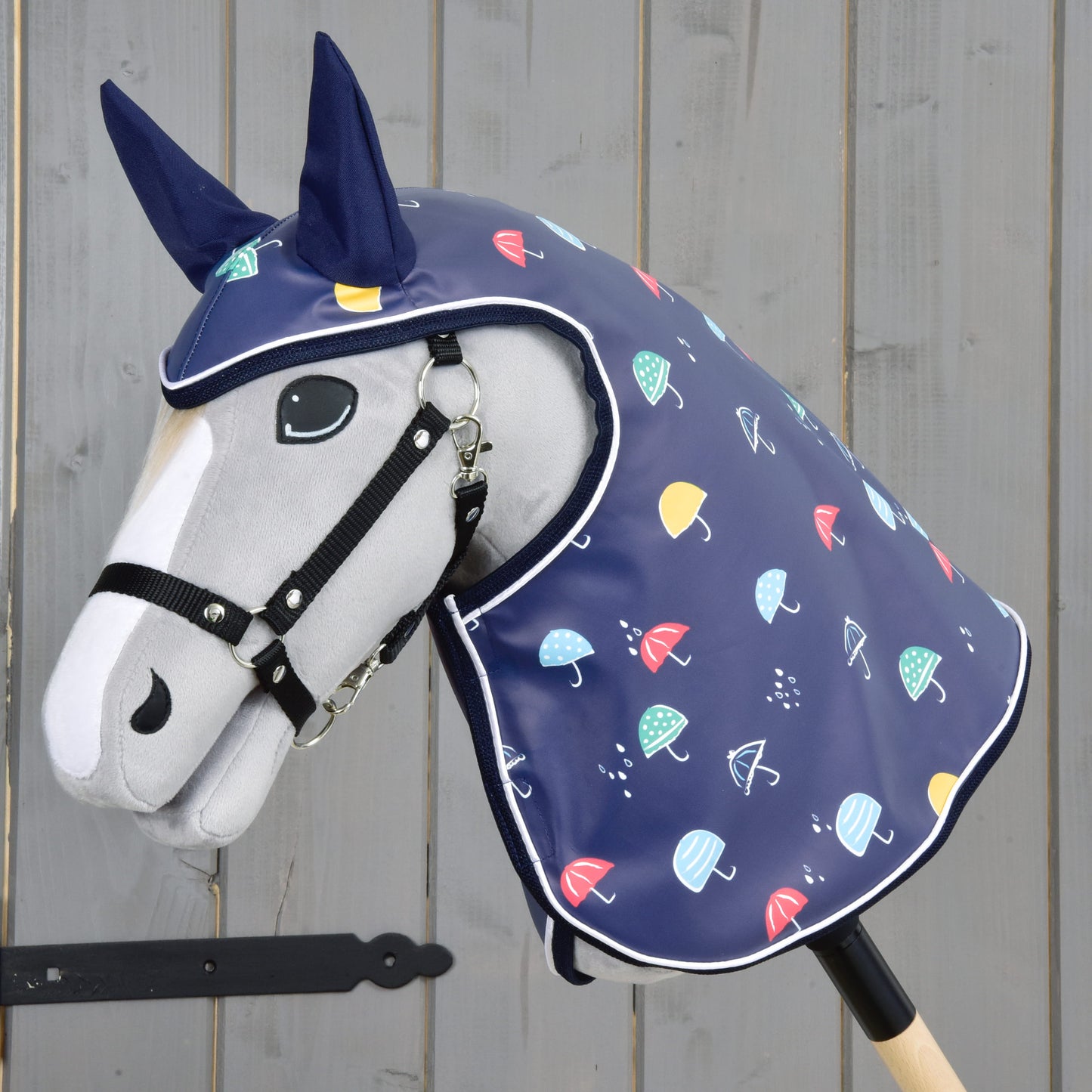 Hobby Horse LarDen „Pláštěnka Rainy“ je plyšový Hobby Horse s koňskou hlavou na dřevěné tyči, který má na sobě modrou pláštěnkovou kapuci do každého počasí s barevným vzorem deštníků a tmavě modrými kryty uší, zobrazený u šedé dřevěné stěny.