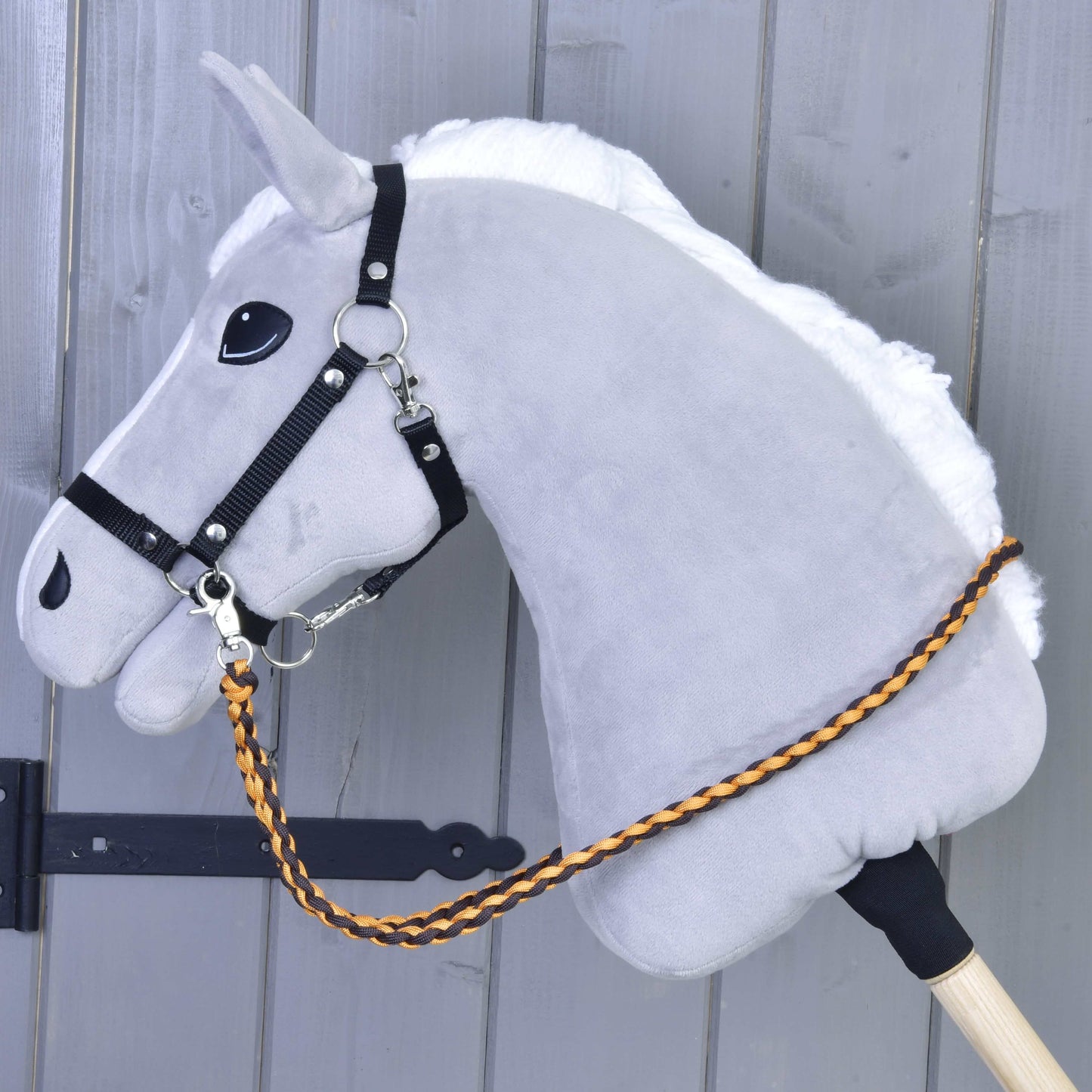 Pletené otěže Knitted Pomello od Hobby Horse LarDen na šedém Hobby Horse s bílou hřívou a černou uzdečkou — pletené polypropylenové otěže v oranžové a hnědé barvě — nasazeném na dřevěné tyči před šedým dřevěným pozadím.