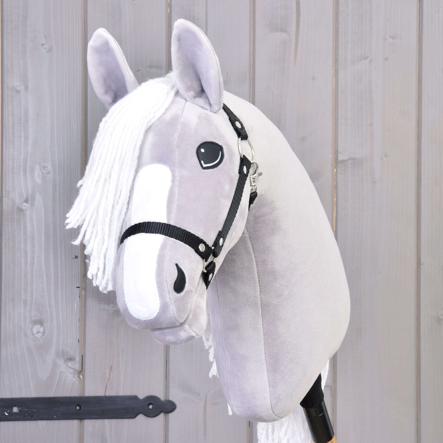 Hobby Horse Popcorn s černou ohlávkou od Hobby Horse LarDen, se šedým plyšovým provedením, bílou hřívou a přátelskou tváří – perfektní společník pro nápaditou hru. Zobrazený před světlou dřevěnou stěnou.