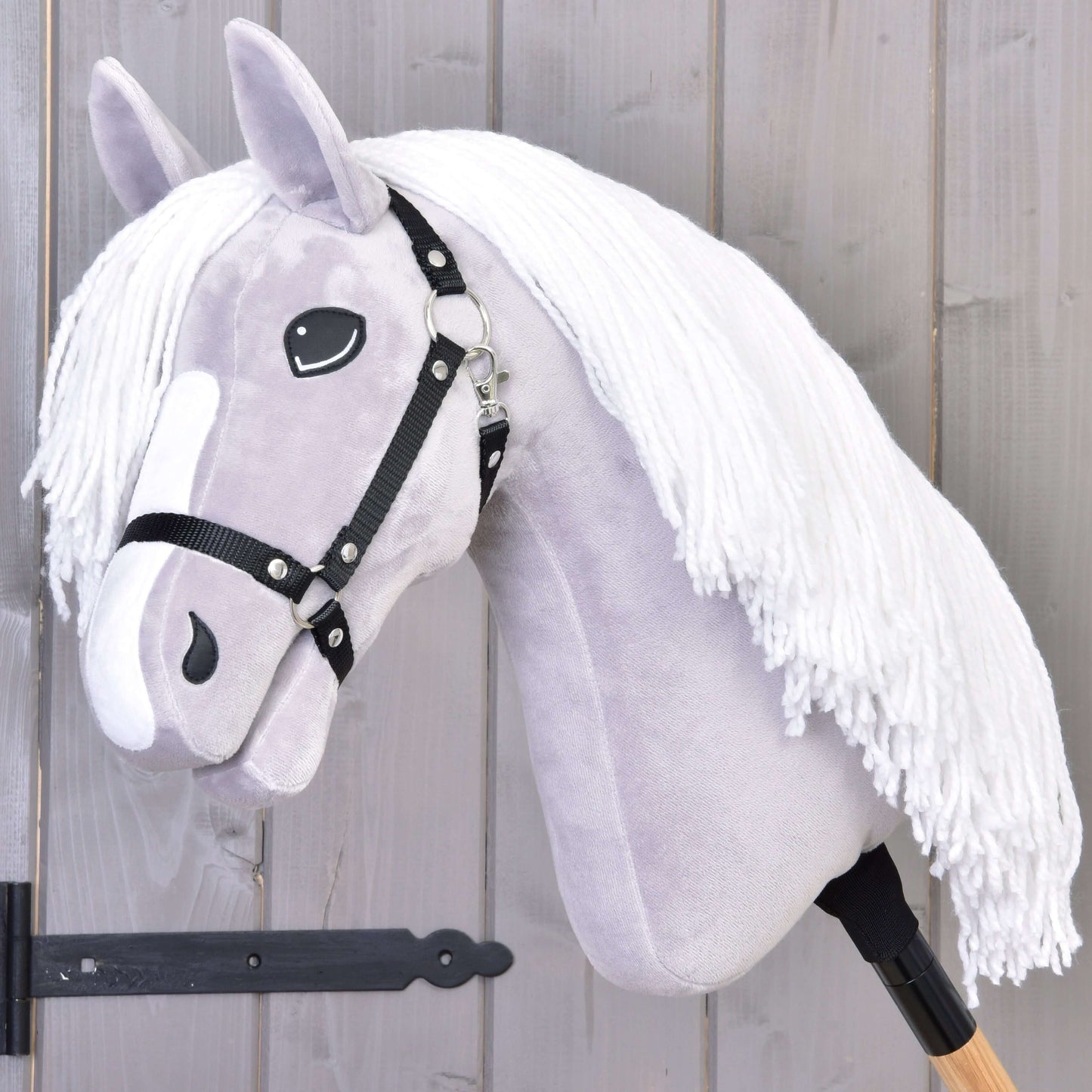Hobby Horse Popcorn s černou ohlávkou od Hobby Horse LarDen, se světle šedou hlavou, bílou přízovou hřívou, černou ohlávkou a dřevěnou tyčí. Ideální pro nápaditou hru a rodinná koňská dobrodružství. Zobrazené před dřevěnou stěnou.