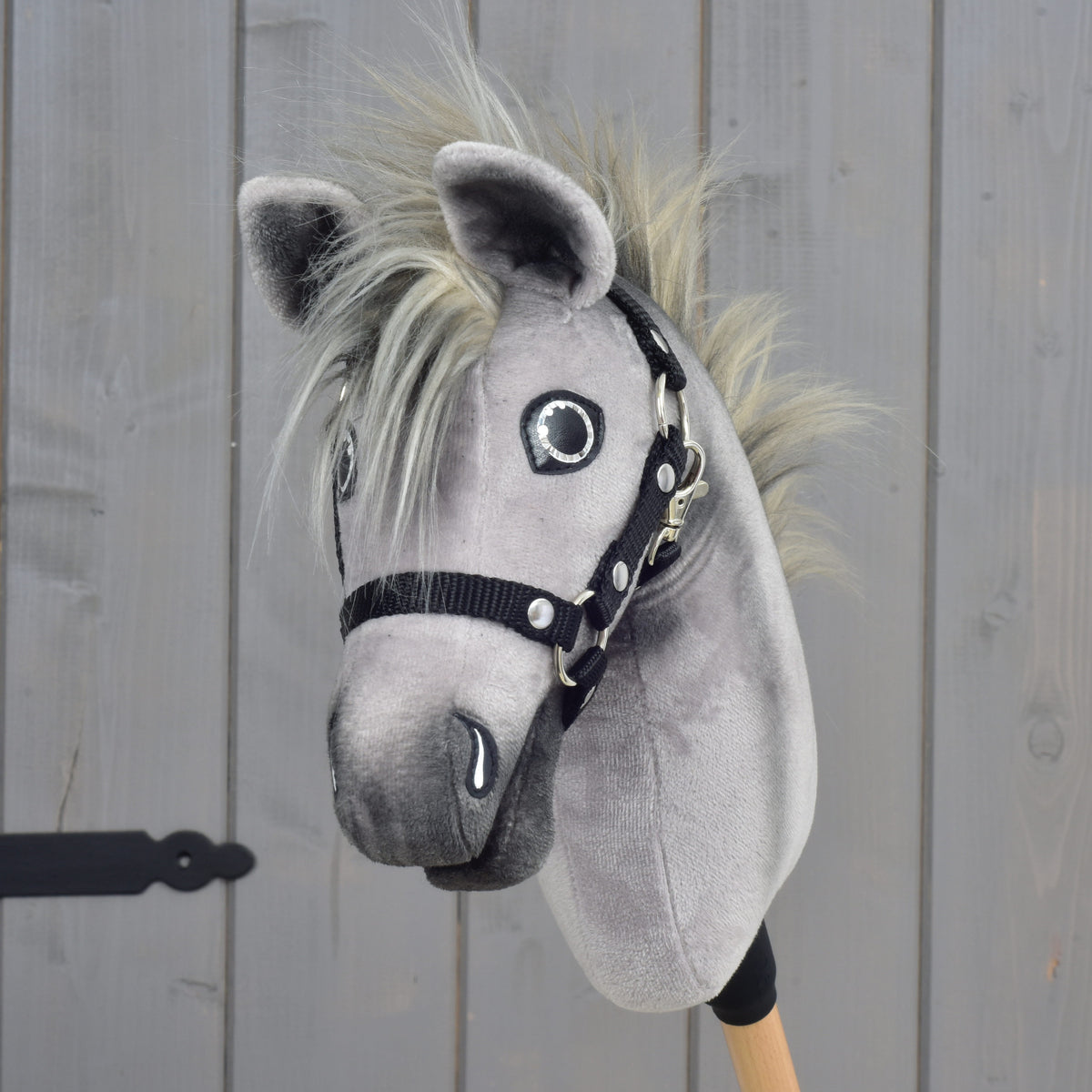 Hobby Horse Prestige s černou ohlávkou, velikost XS od Hobby Horse LarDen, se šedým plyšovým tělem, blond hřívou a na dřevěné tyči, vystavený před světle hnědou dřevěnou stěnou.