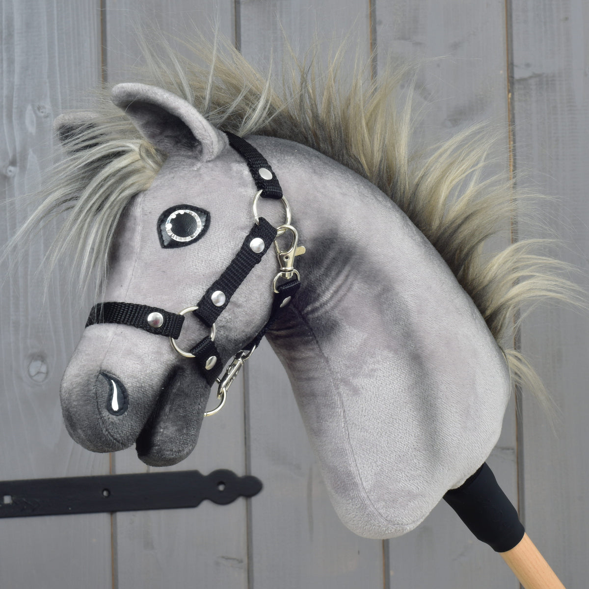 Hobby Horse Prestige s černou ohlávkou, velikost XS od Hobby Horse LarDen, šedý plyšový model se stojatou hřívou a černou ohlávkou, inspirovaný elegancí anglického teplokrevníka. Je nasazený na dřevěné tyči a zobrazený na pozadí dřevěných panelů.