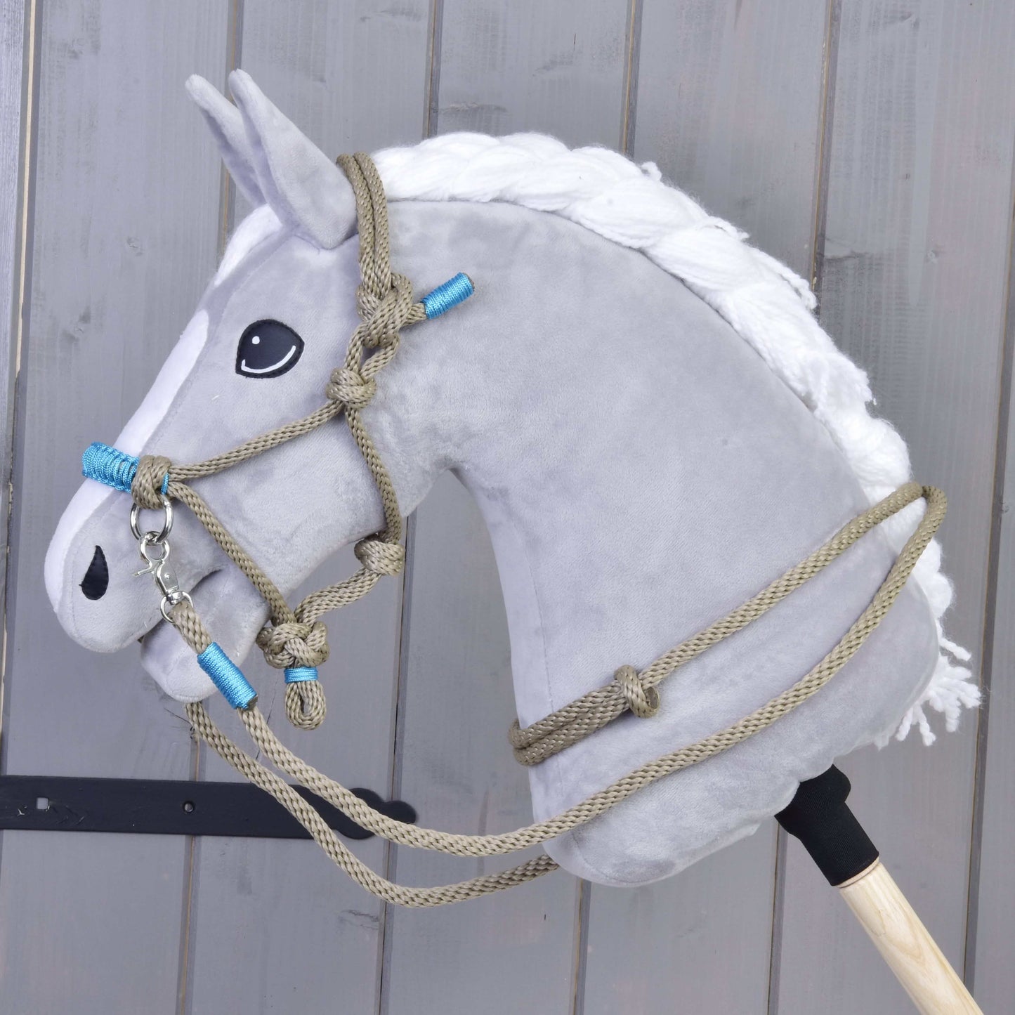 Plyšová šedá hlava Hobby Horse LarDen s bílou hřívou a ohlávkou Provazový set Nature Blue je nasazená na dřevěné tyči a umístěná před pozadím ze šedých dřevěných panelů.