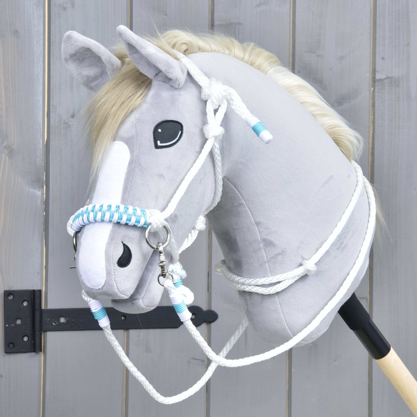 Hobby Horse LarDen Provazový set White Queen s plyšovou hlavou Hobby Horse ve světle šedé barvě, bílou hřívou a pruhem na obličeji a modrobíle pletenými otěžemi, na pozadí dřevěného plotu.