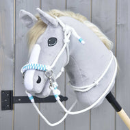 Hobby Horse LarDen Provazový set White Queen s plyšovou hlavou Hobby Horse ve světle šedé barvě, bílou hřívou a pruhem na obličeji a modrobíle pletenými otěžemi, na pozadí dřevěného plotu.