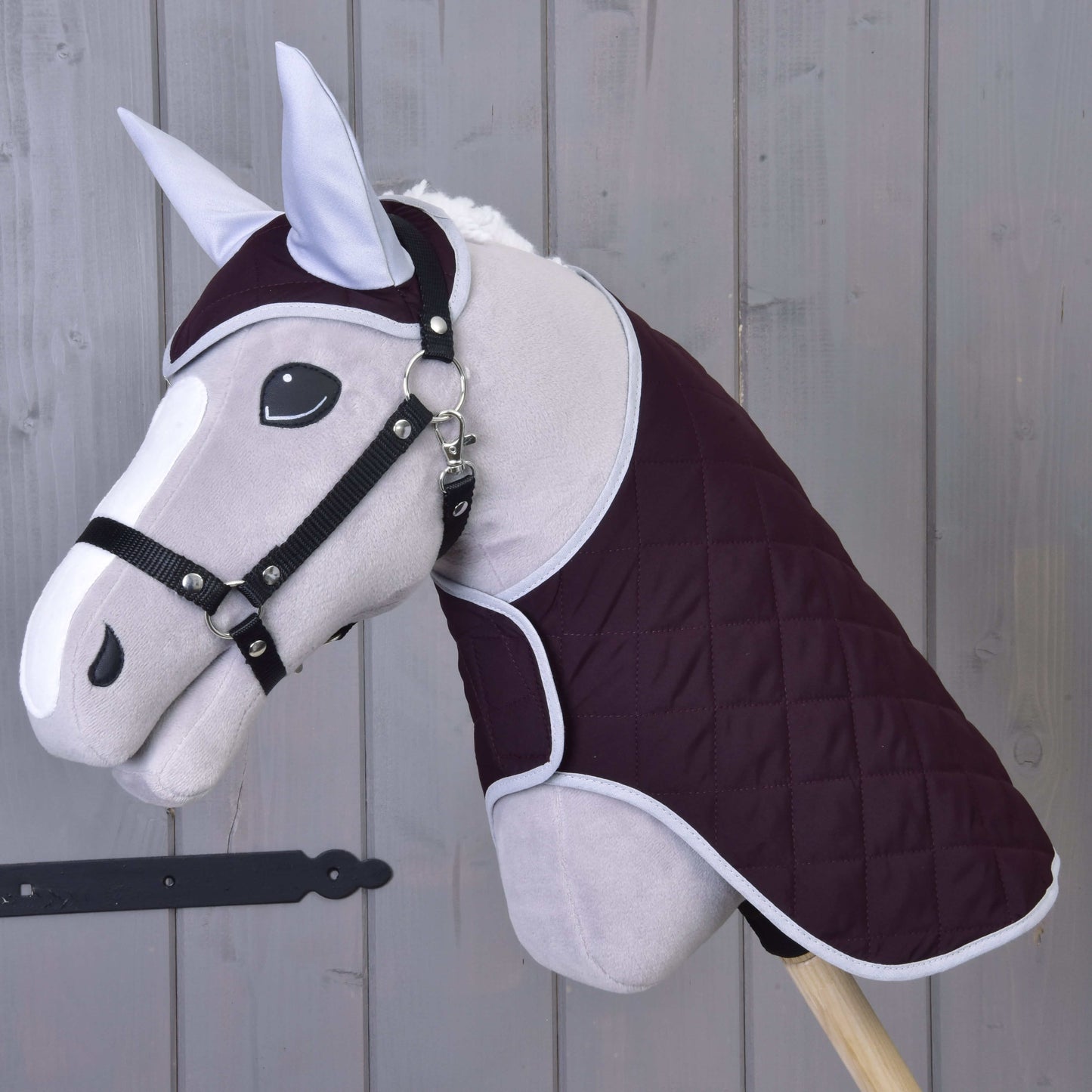 Hobby Horse LarDen Set Purple obsahuje šedou hračku Hobby Horse s prošívanou tmavě fialovou dekou, uzdečkou a náušníkem, vše nasazené na dřevěné tyči a zobrazené na pozadí ze dřevěných prken.