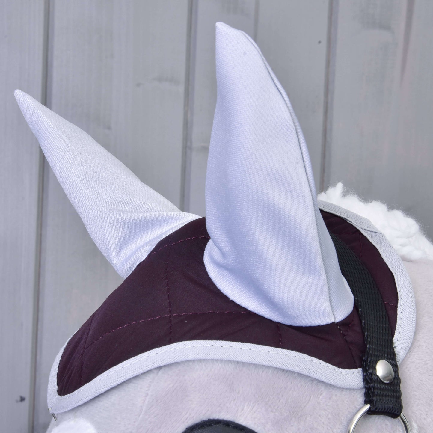 Detail Set Purple od Hobby Horse LarDen: plyšová koňská hlava s velkými šedými oušky, vínovo-šedou prošívanou uzdečkou a náušníkem. V pozadí jsou svislé panely ze světle šedého dřeva.