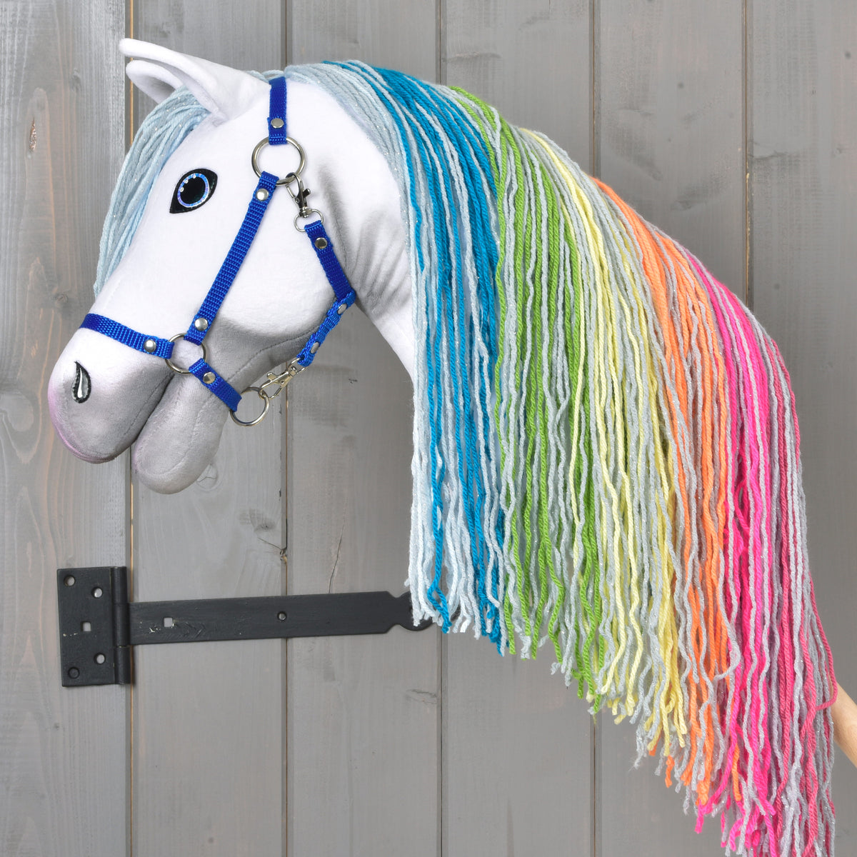 Hobby Horse Rainbow New od LarDen s modrou uzdečkou a dlouhou duhovou hřívou z příze visí na nástěnném držáku před dřevěným pozadím a dodává pokoji pohádkové kouzlo