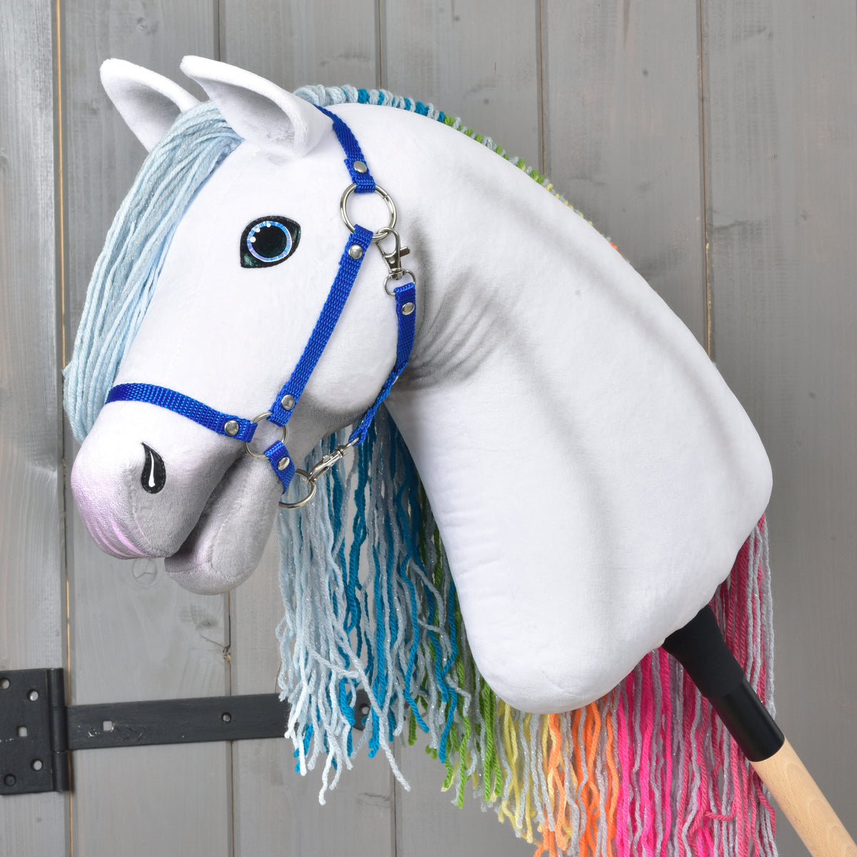 Hravý Hobby Horse Rainbow New od LarDen s modrými otěžemi, světle modrou hřívou z příze a pohádkovou duhovou hřívou na ocase na pozadí šedého dřeva