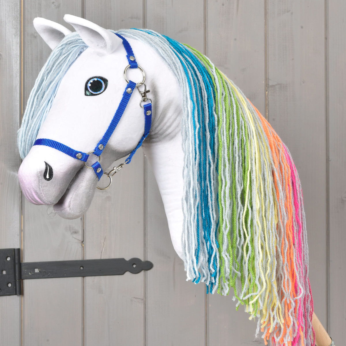 Hobby Horse Rainbow New od LarDen má bílé tělo, dlouhou duhovou hřívu z příze a modrou ohlávku pro pohádkový vzhled na pozadí šedého dřevěného obložení s černým kovovým pantem vlevo