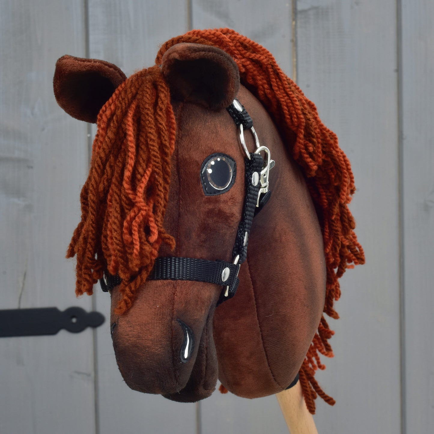 Hobby Horse Rebel s černou ohlávkou - velikost XS od Hobby Horse LarDen, hnědý látkový model navržený tak, aby připomínal hannoverského hřebce, s oranžovou přízovou hřívou, vystavený před šedou dřevěnou stěnou.